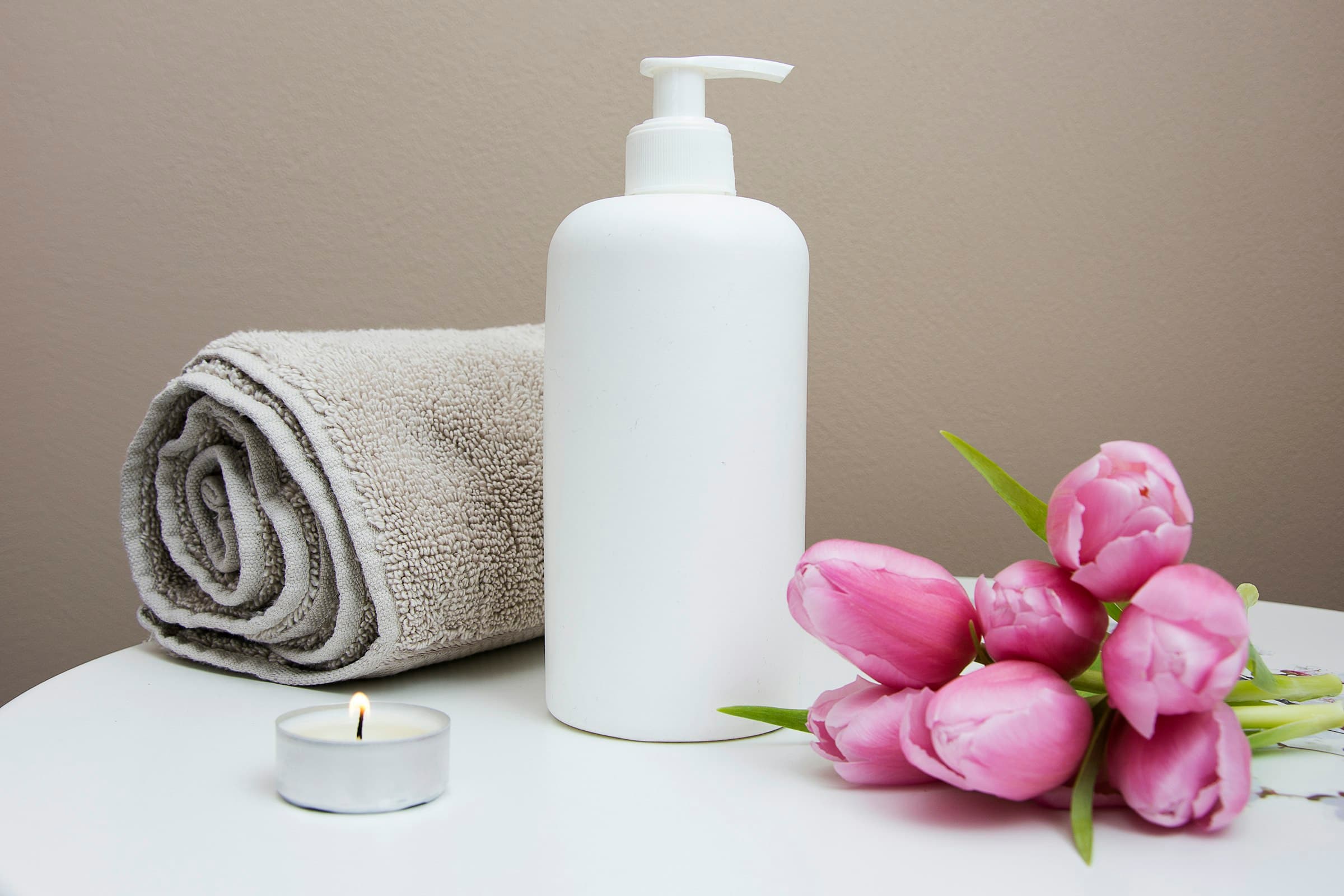 Rolled spa towel, lit candle, and bath bottle with pink tulips on a white counter