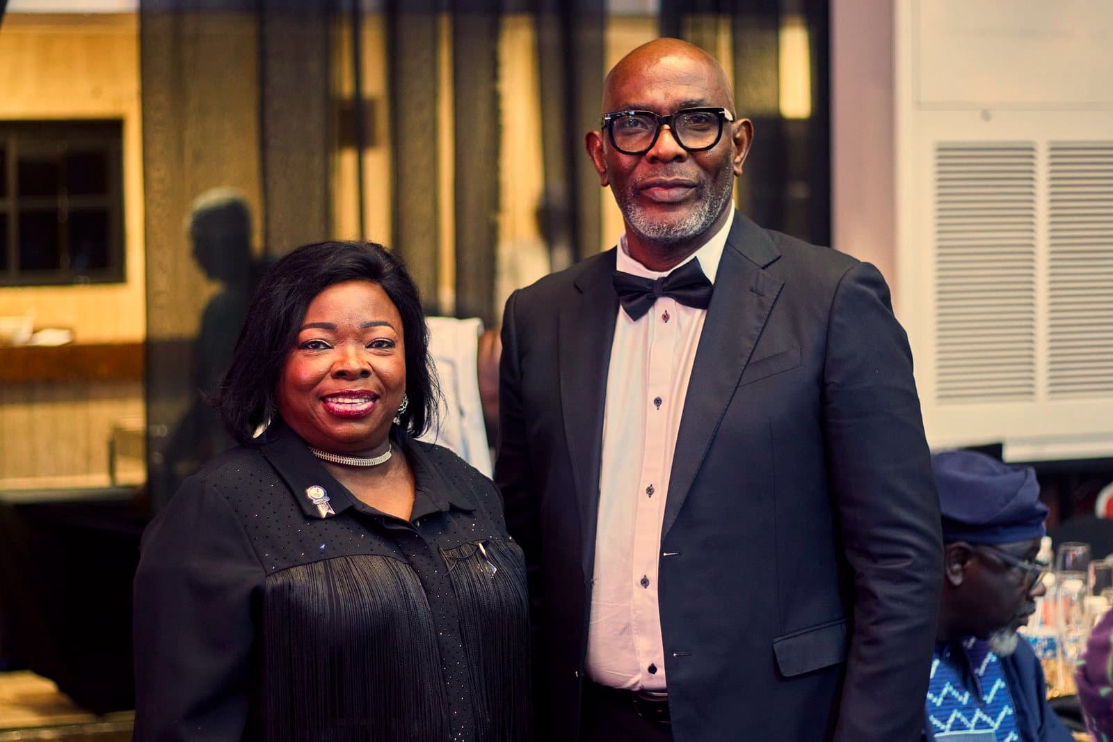 Two elegantly dressed individuals at a black tie event indoors, smiling for the camera.