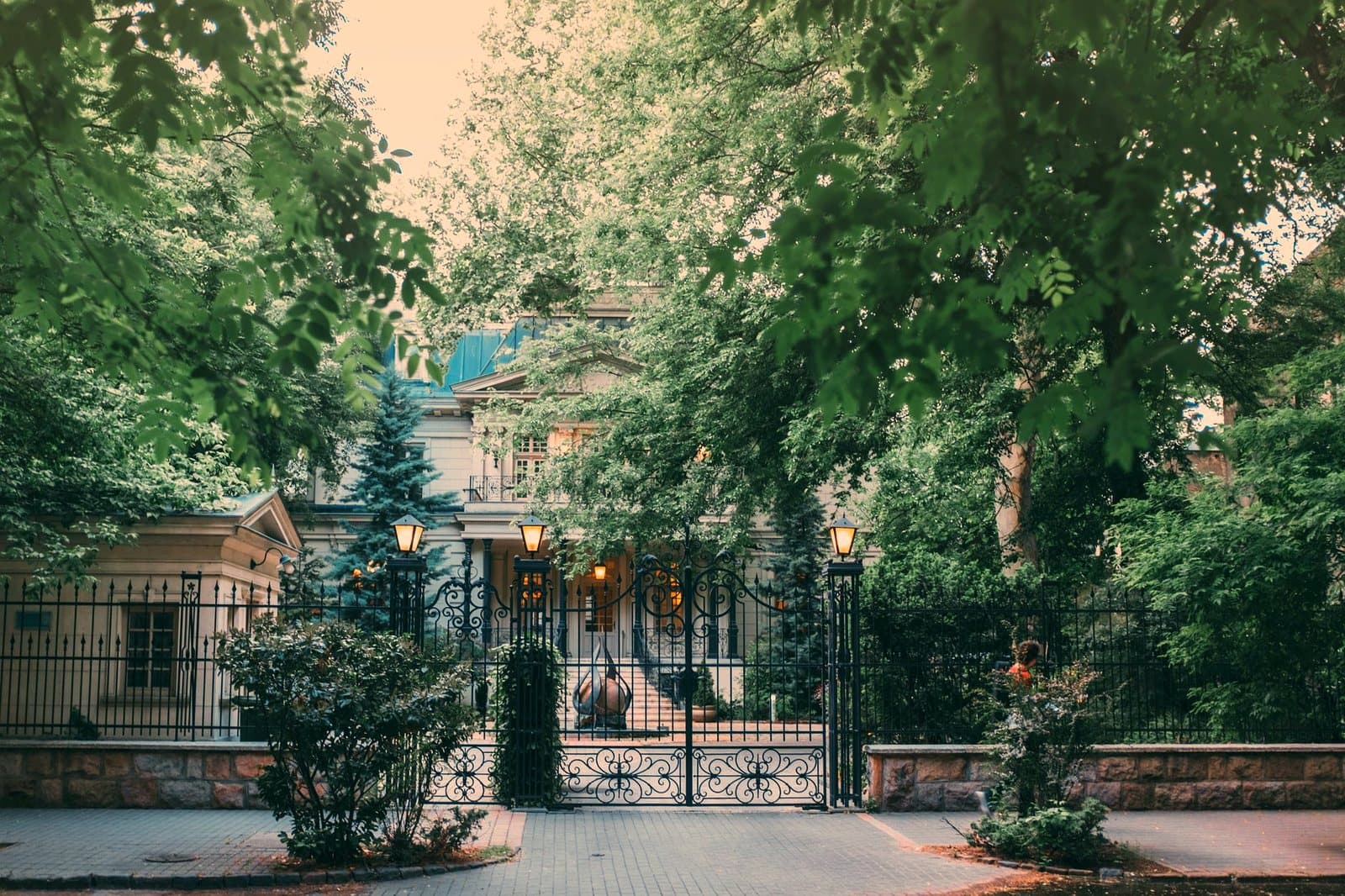 Exterior of stylish classic styled mansion with forged fence and elegant street lamps surrounded by lush green trees in