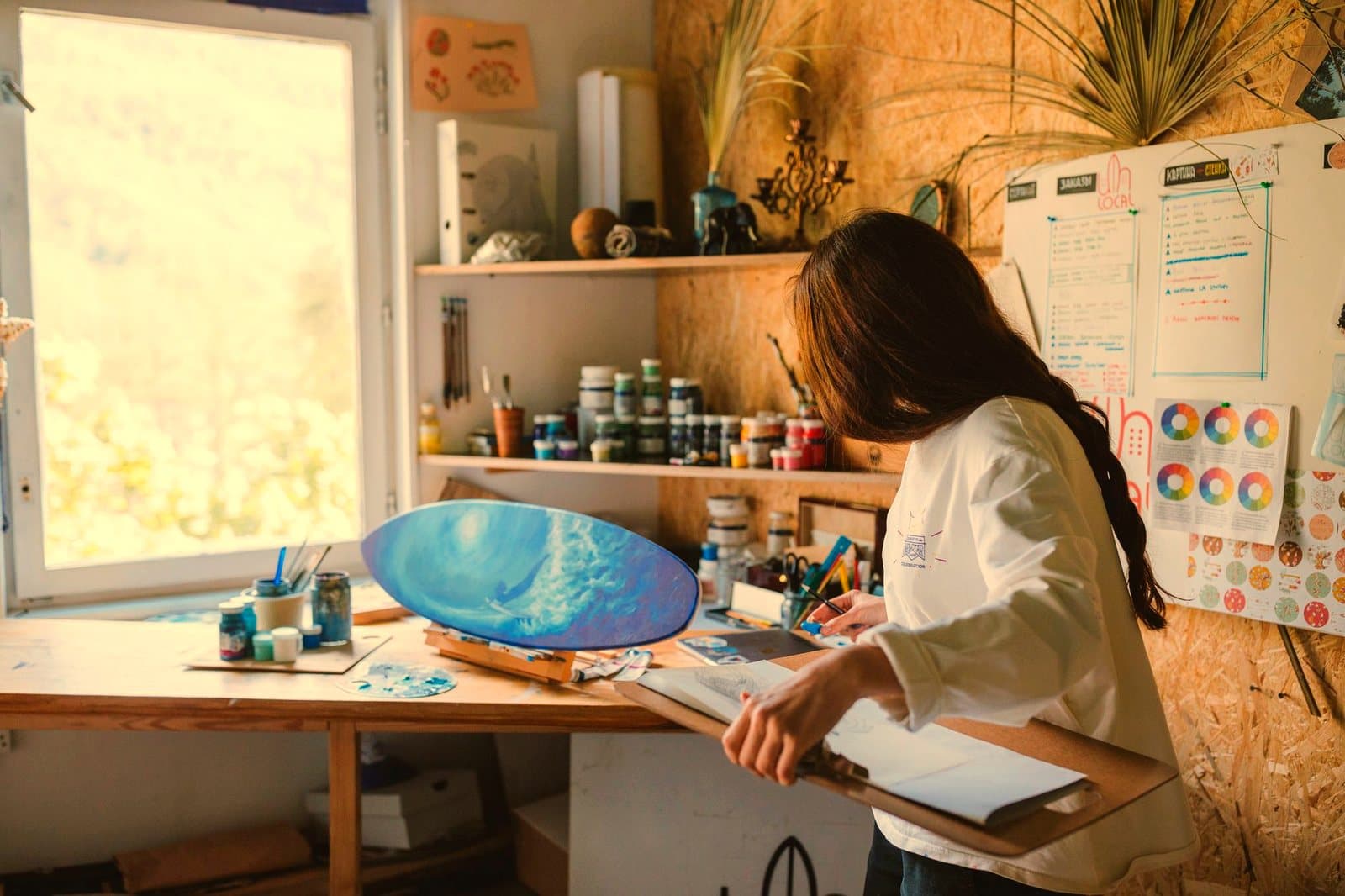 Woman artist painting a blue ocean scene on a skateboard in a cozy art studio.